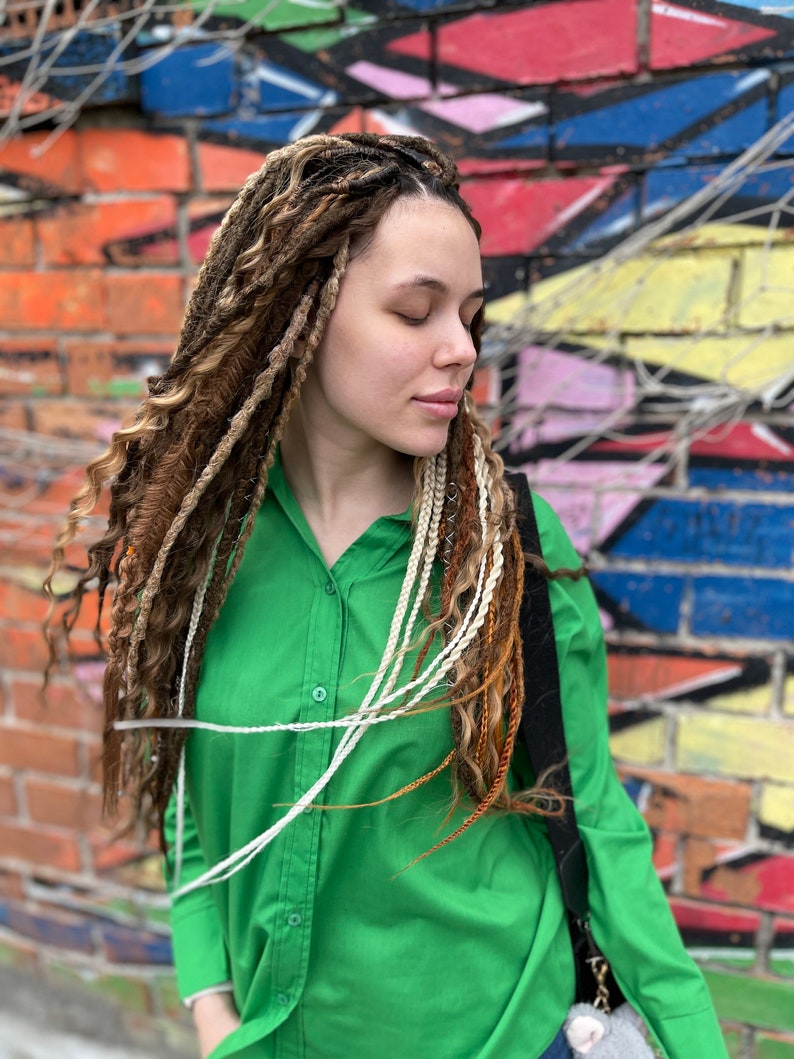 Pode incluir: Uma mulher com tran&ccedil;as longas, castanhas e loiras, usando uma camisa verde com bot&otilde;es. Ela est&aacute; em p&eacute; em frente a uma parede de tijolos com grafites coloridos.