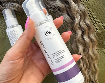 Op de afbeelding: Een witte fles Clear Braids leave-in crème conditioner voor krullend haar. De fles heeft een pompdosser en een paarse en roze band. De tekst "CLEAR BRAIDS" staat verticaal op de fles.