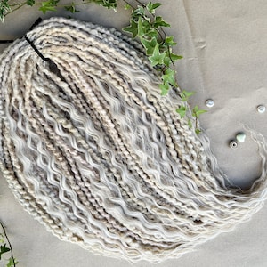 May include: A set of long, wavy, synthetic dreadlocks in a light blonde and white colour. The dreadlocks are arranged in a loose, flowing style.