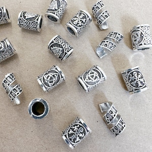 May include: A collection of silver-toned metal beads with various designs, including Celtic knots, snowflakes, and geometric patterns. The beads are arranged on a brown surface.