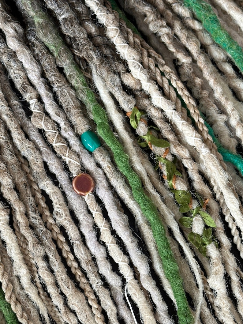 May include: Close-up of a collection of synthetic dreadlocks in shades of beige and green. The dreadlocks are decorated with small beads and fabric flowers.