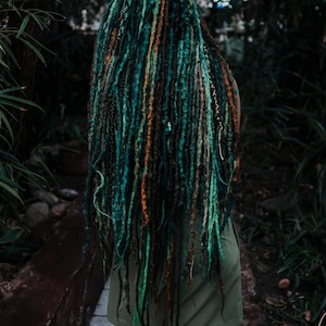 Pode incluir: Uma pessoa com longos dreadlocks verde-azulados, verdes e castanhos. Os dreadlocks s&atilde;o estilizados em uma variedade de cores e texturas. A pessoa est&aacute; vestindo um vestido verde sem mangas.