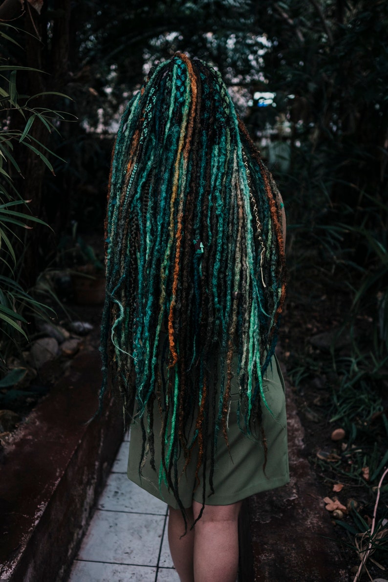 Pode incluir: Uma pessoa com longos dreadlocks verde-azulados, verdes e castanhos. Os dreadlocks s&atilde;o estilizados em uma variedade de cores e texturas. A pessoa est&aacute; vestindo um vestido verde sem mangas.