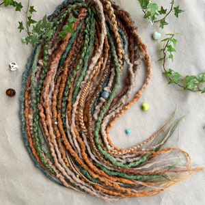 May include: Handmade dreadlocks in a mix of earthy colours: greens, browns, and beige. The dreads are decorated with beads and gold thread, giving a bohemian look. The arrangement is styled with ivy and small heart-shaped stones, creating a natural aesthetic.