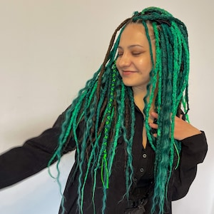 Pode incluir: Uma pessoa com dreadlocks longos, verdes e marrons. Os dreadlocks s&atilde;o decorados com fio verde e azul. A pessoa est&aacute; vestindo uma camisa preta e cal&ccedil;as pretas.