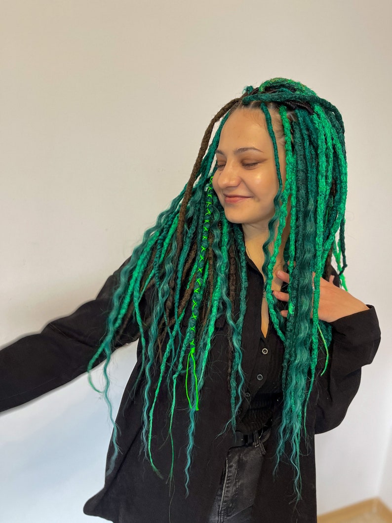 Pode incluir: Uma pessoa com dreadlocks longos, verdes e marrons. Os dreadlocks s&atilde;o decorados com fio verde e azul. A pessoa est&aacute; vestindo uma camisa preta e cal&ccedil;as pretas.