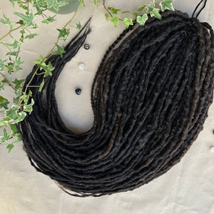 Puede incluir: Un grueso paquete de rastas negras, probablemente de pelo sintético, dispuestas en forma circular. Las rastas están estrechamente tejidas y tienen una apariencia ligeramente texturizada.