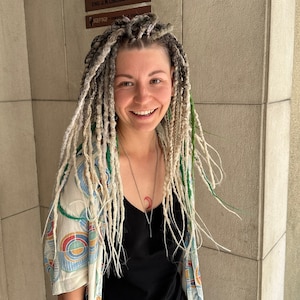 May include: A woman with long, white and green dreadlocks is wearing a black tank top and a white and blue patterned shirt. She is smiling and looking at the camera.