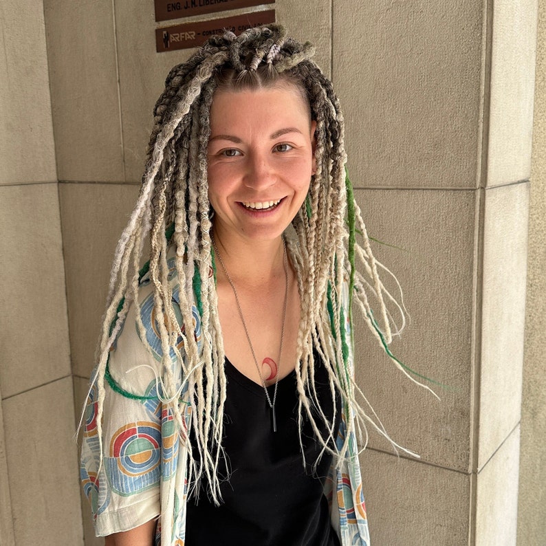 May include: A woman with long, white and green dreadlocks is wearing a black tank top and a white and blue patterned shirt. She is smiling and looking at the camera.