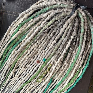 May include: A close-up of a set of synthetic dreadlocks in shades of white, gray, and green. The dreadlocks are tied together with a black ribbon and have beads and other decorative elements.