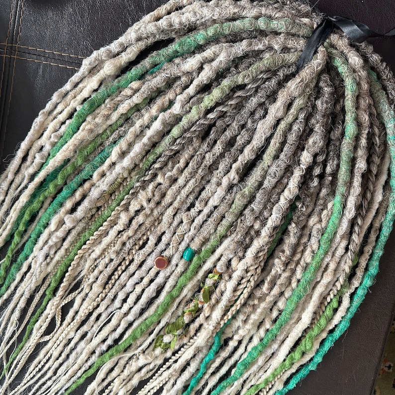 May include: A close-up of a set of synthetic dreadlocks in shades of white, gray, and green. The dreadlocks are tied together with a black ribbon and have beads and other decorative elements.