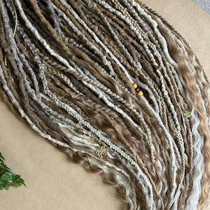 May include: A collection of handmade dreadlocks in various shades of brown, gray, and blonde. The dreads are adorned with wooden beads and small gold-colored charms. Some dreads are braided, while others have a wavy texture.