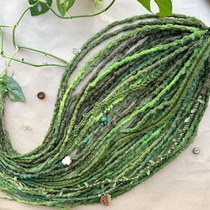 May include: A collection of green dreadlocks in various shades, including olive, lime, and forest green. The dreadlocks are adorned with decorative beads and charms, and a leaf-shaped charm. The dreadlocks are arranged on a neutral-toned surface.