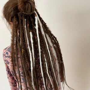Pode incluir: Dreadlocks marrons e brancas com contas e pingentes. As dreadlocks est&atilde;o penteadas em um coque no topo da cabe&ccedil;a.