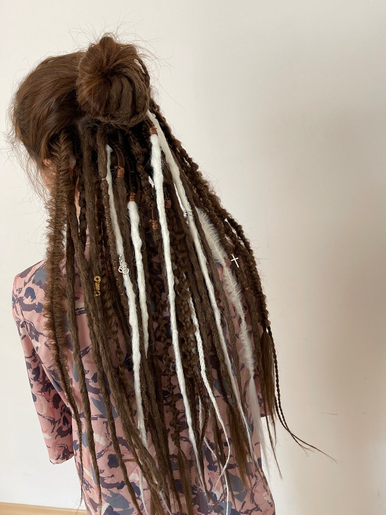 Pode incluir: Dreadlocks marrons e brancas com contas e pingentes. As dreadlocks est&atilde;o penteadas em um coque no topo da cabe&ccedil;a.