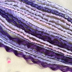 May include: A collection of purple, lavender, and pink synthetic dreadlocks with silver beads. The dreadlocks are arranged in a row and are ready to be installed.