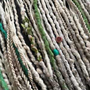 May include: A collection of synthetic dreadlocks in shades of white and green. The dreadlocks are arranged in rows and feature decorative beads and charms.