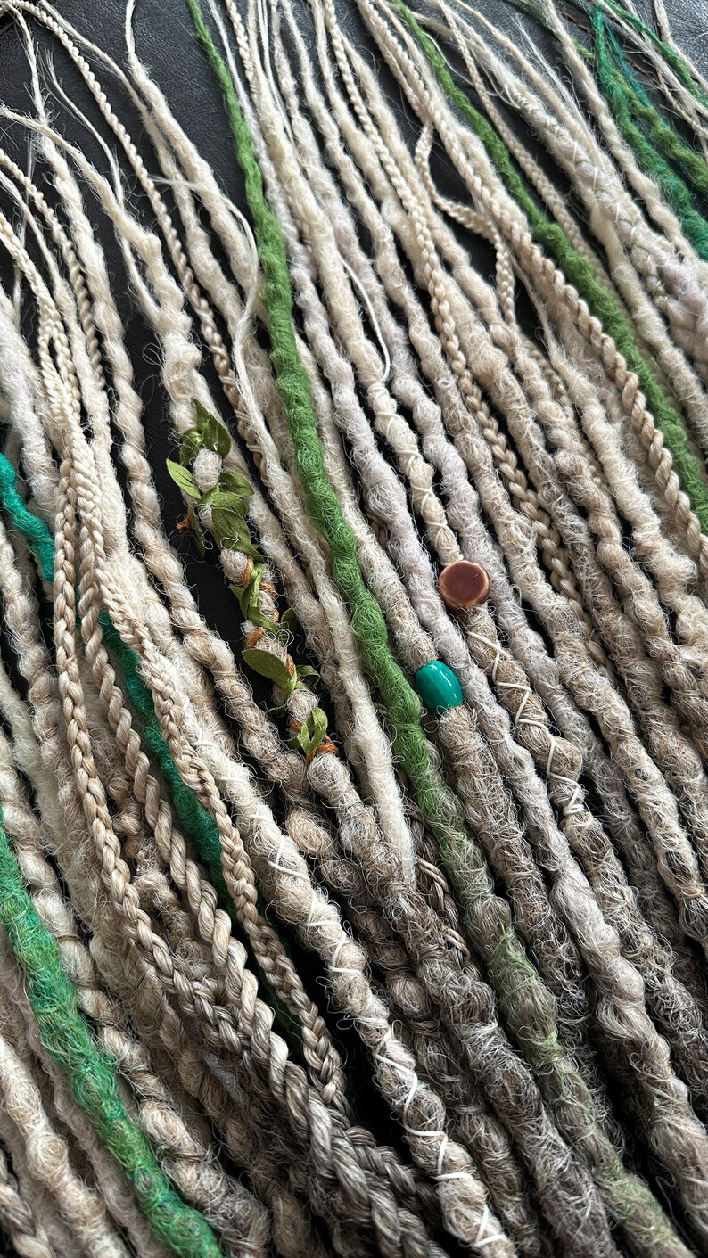 May include: A collection of synthetic dreadlocks in shades of white and green. The dreadlocks are arranged in rows and feature decorative beads and charms.