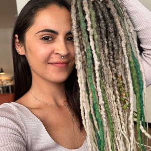 May include: A person with long, dark hair is wearing a light gray sweater. They are holding up a set of synthetic dreadlocks in shades of green, white, and gray.