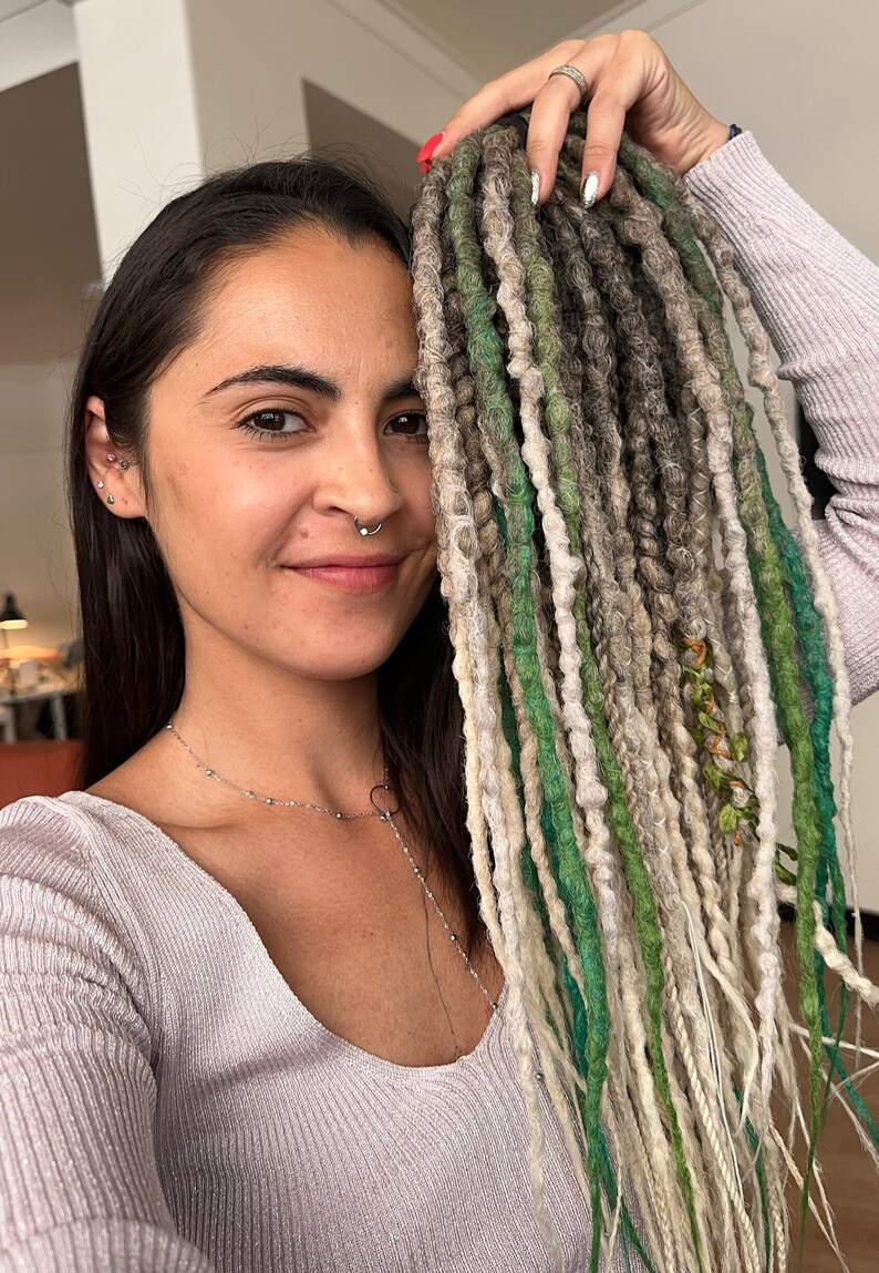 May include: A person with long, dark hair is wearing a light gray sweater. They are holding up a set of synthetic dreadlocks in shades of green, white, and gray.