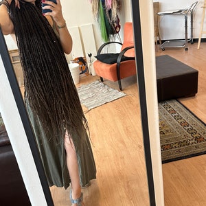 Pode incluir: Uma pessoa com longos dreadlocks marrons est&aacute; em p&eacute; em frente a um espelho vestindo um vestido verde e sand&aacute;lias azuis. A pessoa est&aacute; tirando uma selfie com um smartphone preto.
