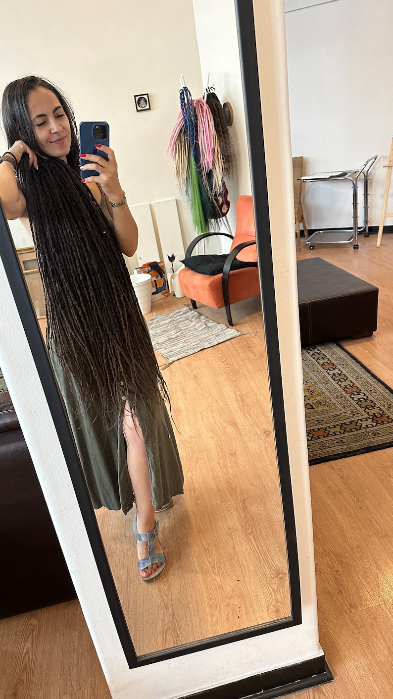Pode incluir: Uma pessoa com longos dreadlocks marrons est&aacute; em p&eacute; em frente a um espelho vestindo um vestido verde e sand&aacute;lias azuis. A pessoa est&aacute; tirando uma selfie com um smartphone preto.