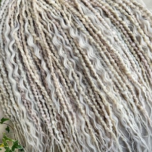 Puede incluir: Primer plano de un paquete de largas rastas sintéticas onduladas, de color rubio claro y blanco. Las rastas están hechas de un material suave y texturizado y tienen un aspecto natural y bohemio.