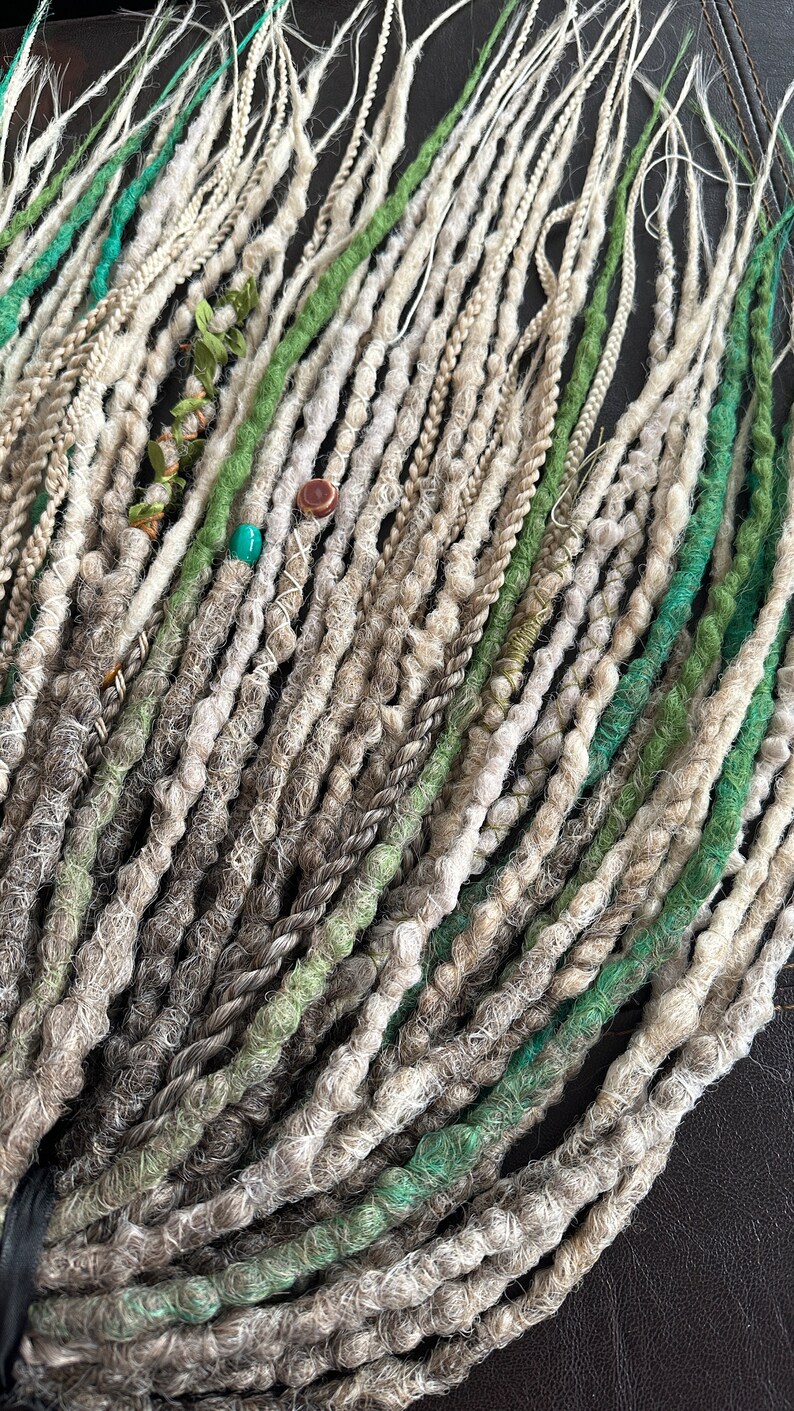 May include: A collection of synthetic dreadlocks in shades of gray, white, and green. The dreadlocks are arranged in a fan shape and have beads and other decorative elements.