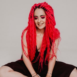 Pode incluir: Uma mulher com longas tran&ccedil;as rosa brilhantes senta-se com as pernas cruzadas, vestindo um vestido vinho escuro. Ela est&aacute; sorrindo e olhando para baixo.