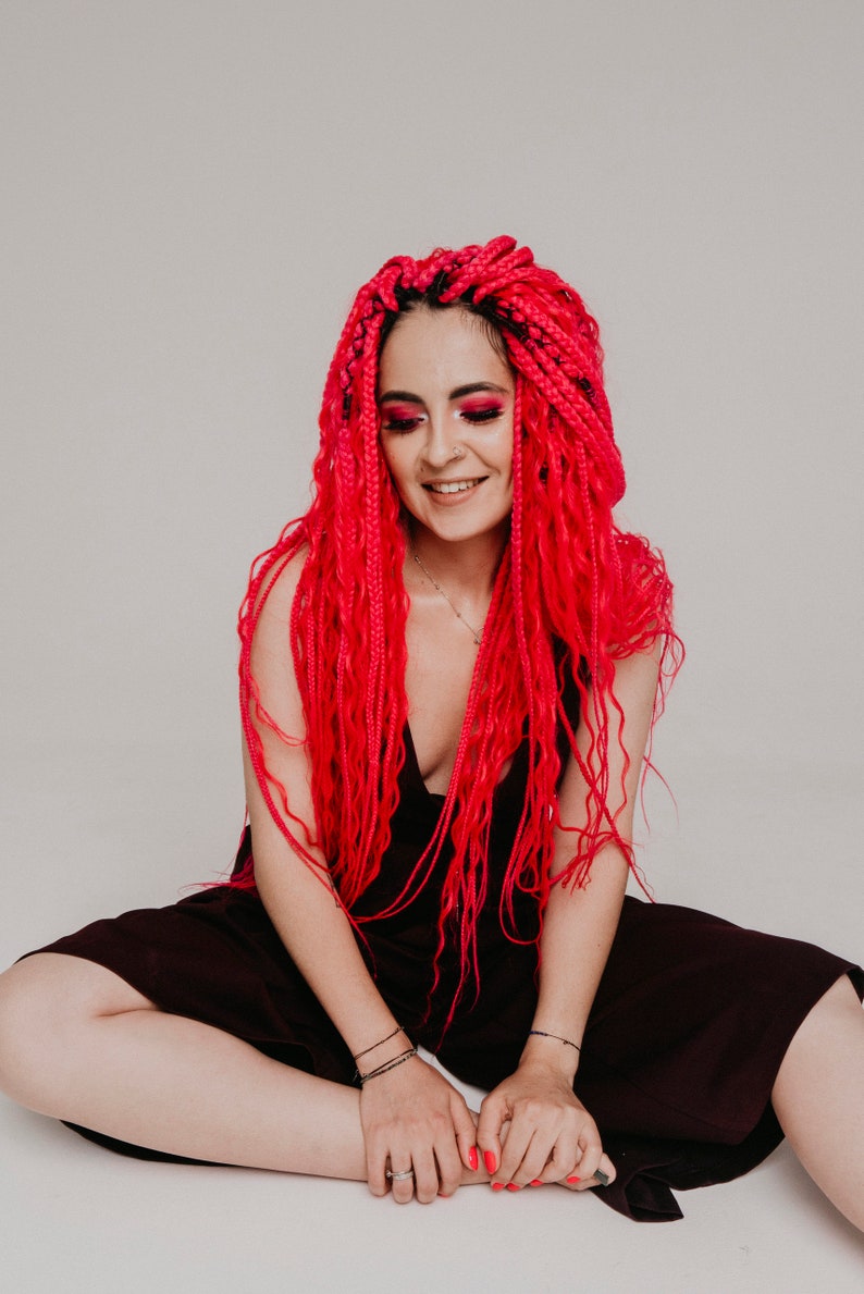 Pode incluir: Uma mulher com longas tran&ccedil;as rosa brilhantes senta-se com as pernas cruzadas, vestindo um vestido vinho escuro. Ela est&aacute; sorrindo e olhando para baixo.