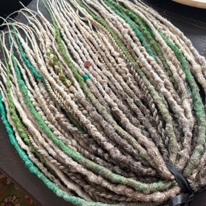 May include: A bundle of synthetic dreadlocks in shades of gray, white, and green. The dreadlocks are tied together with a black ribbon.