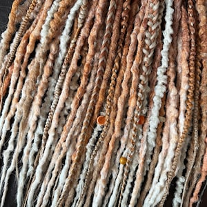 Può includere: Una collezione di dreadlock sintetiche marroni, bionde e bianche con perline. Le dreadlock sono disposte in un fascio e sono pronte per essere installate.