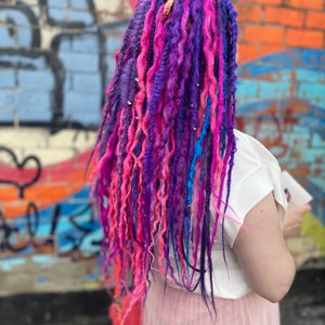 Pode incluir: Uma pessoa com longos dreadlocks coloridos em tons de rosa, roxo e azul. Os dreadlocks s&atilde;o estilizados com contas e s&atilde;o usados ​​nas costas. A pessoa est&aacute; vestindo uma camiseta branca e uma saia rosa.