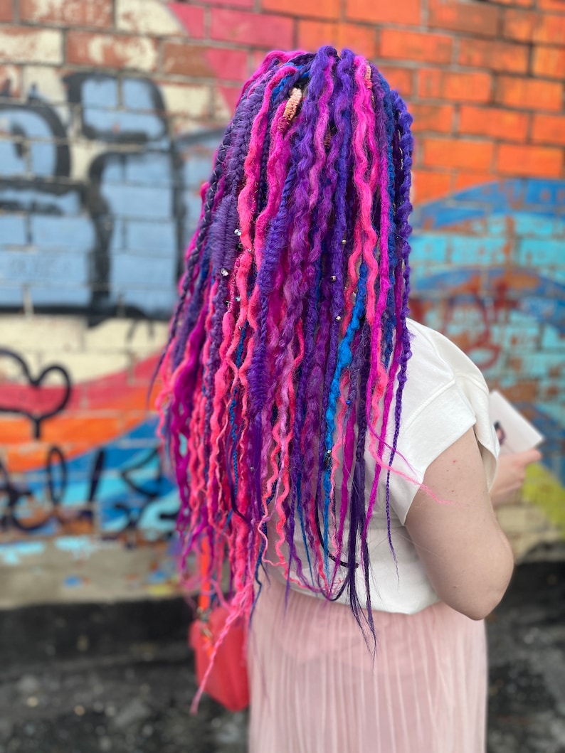 Pode incluir: Uma pessoa com longos dreadlocks coloridos em tons de rosa, roxo e azul. Os dreadlocks s&atilde;o estilizados com contas e s&atilde;o usados ​​nas costas. A pessoa est&aacute; vestindo uma camiseta branca e uma saia rosa.