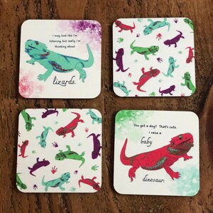 Puede incluir: Cuatro posavasos cuadrados con fondo blanco y diseños de lagartos coloridos. Un posavasos presenta un lagarto verde con el texto "Puedo parecer que estoy escuchando, pero en realidad estoy pensando en lagartos." Otro posavasos presenta un lagarto rojo con el texto "¿Tienes un perro? Es lindo. Crío un dinosaurio bebé."
