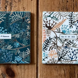 Op de afbeelding: Twee vierkante keramische onderzetters met een botanisch ontwerp. Eén is teal met witte en bruine accenten, de andere is wit met zwarte, bruine en teal accenten. Tekst op de onderzetters luidt "L & P Hanson" en "Est: 15-03-2025".