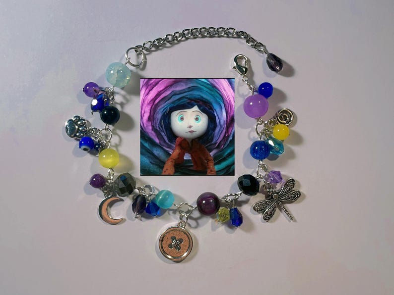 May include: A silver charm bracelet with a variety of colorful beads and charms, including a button, a crescent moon, a dragonfly, and a small picture of a girl with blue eyes. The beads are in shades of blue, purple, yellow, and green.