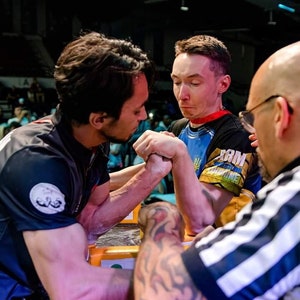 Op de afbeelding: Twee armworstelaars staan tegenover elkaar en grijpen elkaar bij de handen in een wedstrijd. De deelnemers dragen sportshirts, met een scheidsrechter in een gestreept shirt die toeziet. Het evenement vindt binnen plaats, met toeschouwers op de achtergrond.