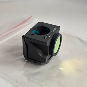 May include: A black, rectangular optical device with a green lens and a blue interior. The device has the text "N 2.1 513 810" printed on its side. The device is sitting on a clear plastic surface.