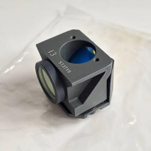 May include: A dark gray optical device with a circular lens and the numbers "13" and "513719" printed on the side. The device has a square shape with a circular opening and a blue interior.