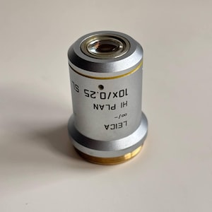 Puede incluir: Objetivo de microscopio plateado y dorado con el texto "LEICA HI PLAN 10x/0.25 SL". El objetivo tiene un cuerpo cilíndrico plateado, una base dorada y una lente transparente.