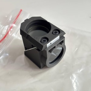 May include: A dark gray, rectangular optical component with two circular openings and a small label marked "A". The component is secured with two screws and appears to be made of a durable material. The item is on a white surface.