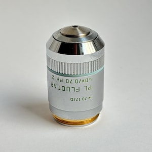 Puede incluir: Objetivo de microscopio plateado y dorado con la inscripción "PL FLUOTAR 40x/0.70 PH 2". Presenta un cuerpo cilíndrico con una sección estriada y una parte superior redondeada.