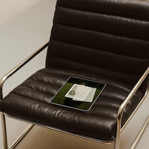 May include: A dark brown leather armchair with a chrome frame. A tablet with a photo is on the seat. The chair has a ribbed design on the back and seat. The background is a neutral color.