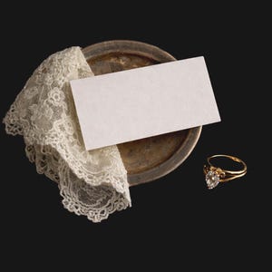 May include: A gold ring with a heart-shaped diamond sits beside a white card and a vintage lace doily on a tarnished metal plate. The ring has a delicate band against a dark background.