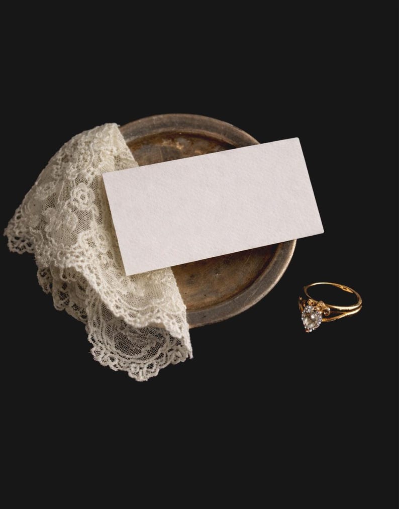 May include: A gold ring with a heart-shaped diamond sits beside a white card and a vintage lace doily on a tarnished metal plate. The ring has a delicate band against a dark background.