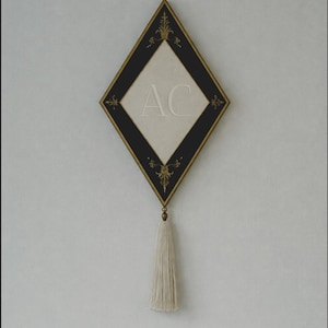 May include: A diamond-shaped wall hanging with a black border and gold accents. The center is off-white with the letters "AC" in the center. A cream-colored tassel hangs from the bottom. Decorative wall art.