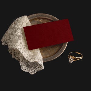 May include: A gold ring with a heart-shaped diamond is placed near a red card and a vintage silver plate with a white lace doily. The ring has a delicate design, and the card has a textured surface. The items are arranged on a dark background.