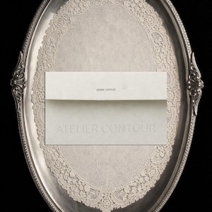 Könnte beinhalten: Ein ovaler, silberfarbener Tablett mit verzierten Rändern hält eine cremefarbene Deckchen und zwei cremefarbene Karten. Auf der oberen Karte steht "atelier contour" und auf der unteren Karte "ATELIER CONTOUR". Der Hintergrund ist schwarz.