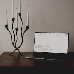Op de afbeelding: Een zwarte metalen kandelaar met zes witte kaarsen staat op een donkere houten tafel. Een laptop toont de tekst "ATELIER CONTO" en een citaat over materiaal en herinnering.